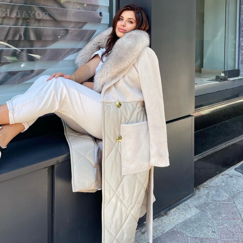 Woman in a long beige coat with fur collar sitting on a bench in an urban setting.