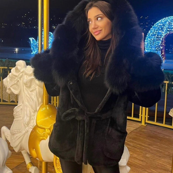 Woman in a dark coat with fur trim standing on a decorated outdoor deck at night.