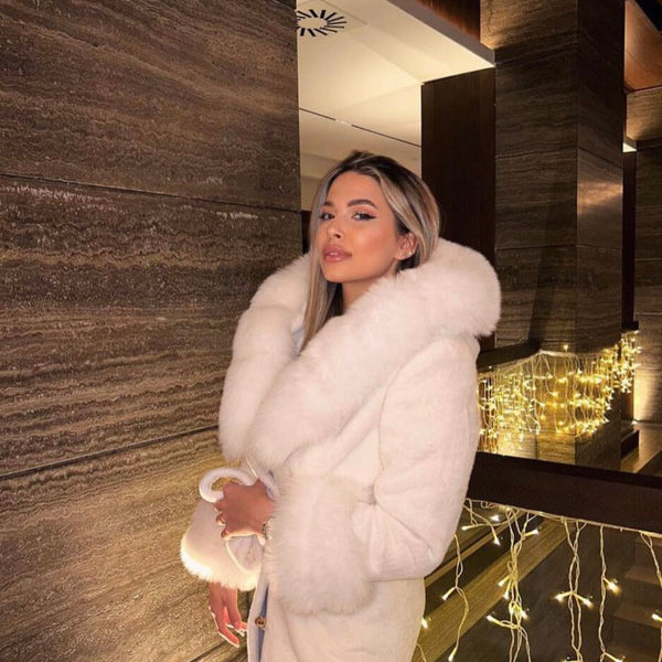 Woman in a white fur coat standing in a modern indoor setting with decorative lights.