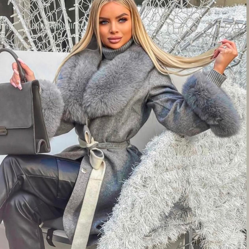 Woman in a gray coat with fur trim holding a handbag, standing against a decorative background.