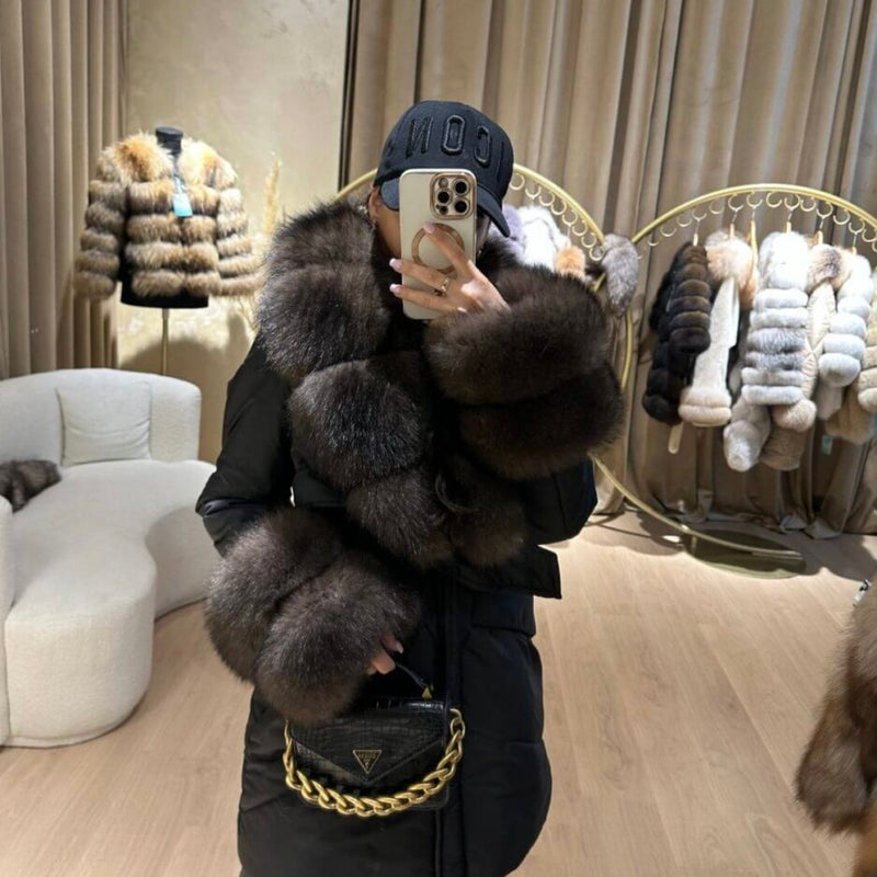 Person wearing a fur vest and taking a mirror selfie in a store with fur coats displayed.