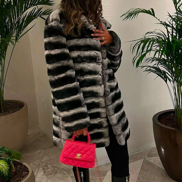 Person wearing a long fur coat holding a pink handbag indoors with plants in the background
