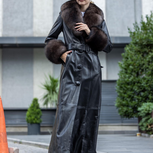 Woman wearing a long black leather coat with fur trim outdoors.