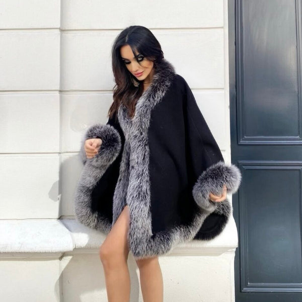 Woman wearing a black poncho with gray fur trim sitting on a bench.