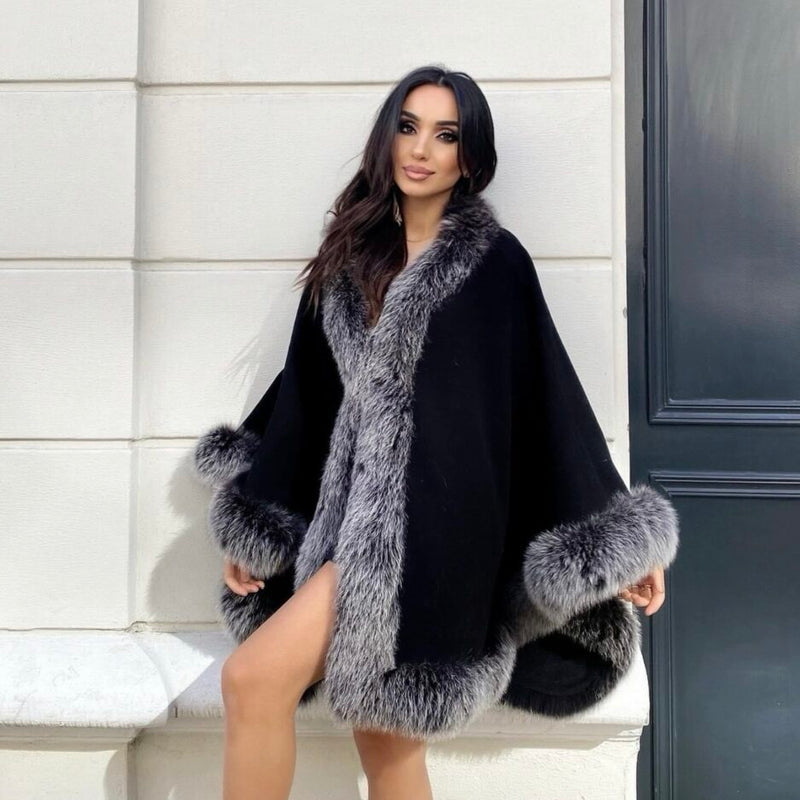 Woman wearing a black cape with gray fur trim sitting on a bench.