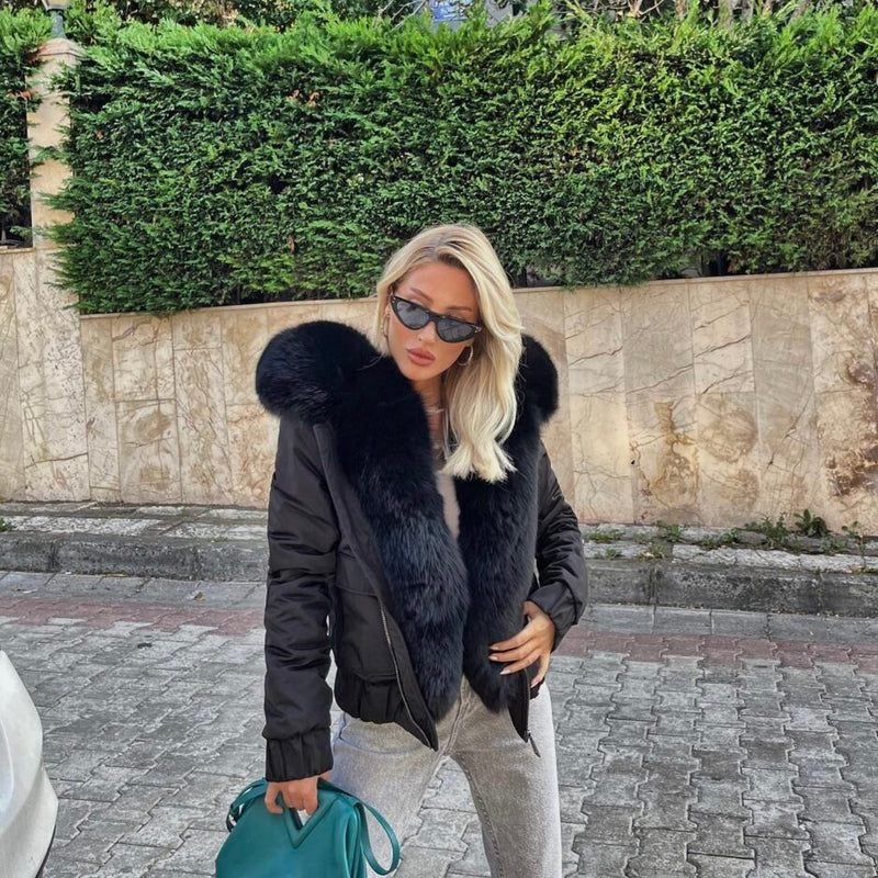 Woman wearing a black fur-lined jacket with sunglasses and a teal handbag on a street.