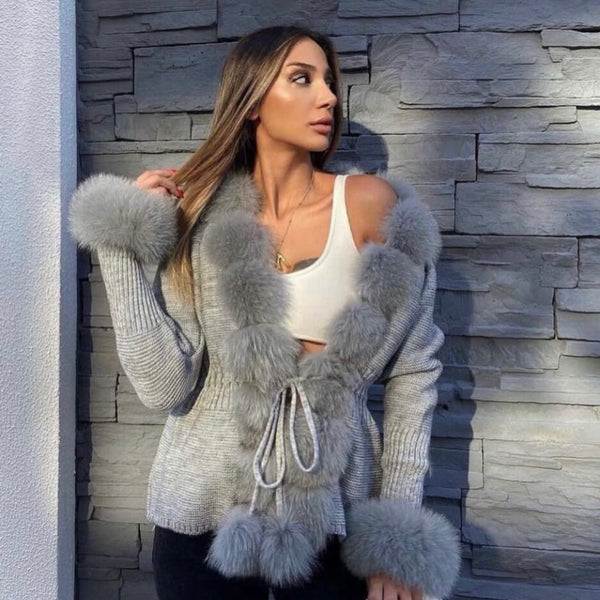 Woman wearing a gray cardigan with fur trim against a stone wall.