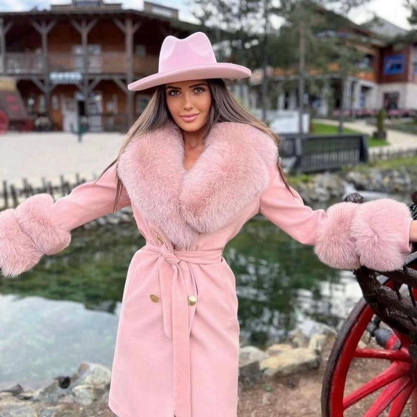 Person in a pink coat and hat standing by a lake with a wooden building in the background