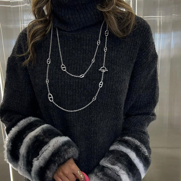 Person wearing a dark sweater with fur-trimmed cuffs, holding a pink handbag, and wearing a silver necklace.